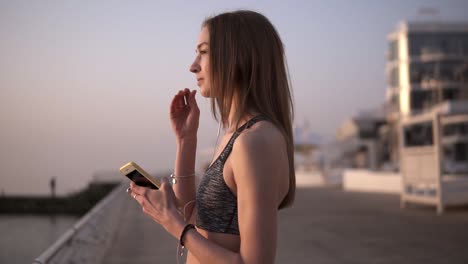 a bright, slender girl stands in the morning near the sea, puts on headphones, a mobile phone in her hands. preparing for training. in a sports bra. overview footage