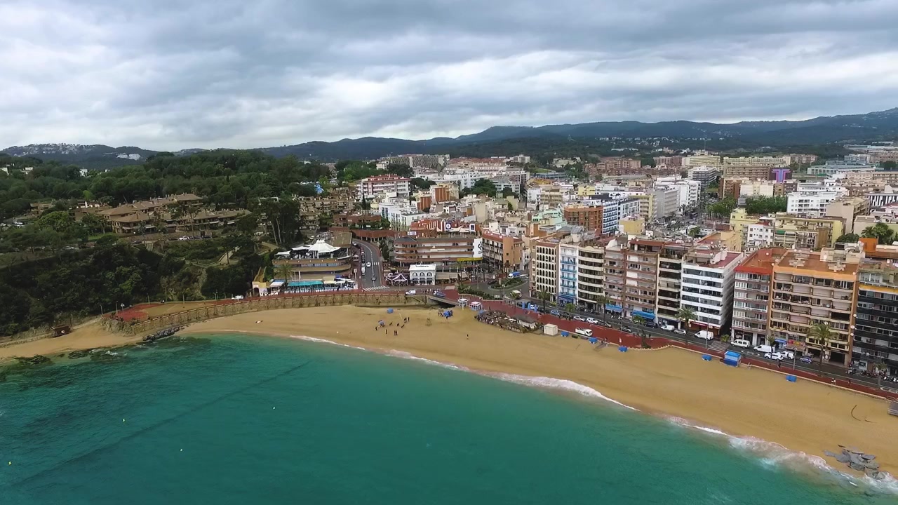 Drone che vola verso la baia di Lloret de Mar, rivelando acque turchesi, spiagge mediterranee e il paesaggio urbano costiero della Costa Brava, Spagna.