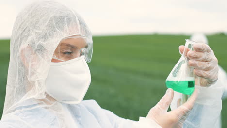 vista de cerca de una investigadora caucásica con traje protector que sostiene un tubo de ensayo mientras realiza el control de plagas en el campo verde