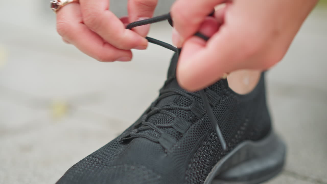 Close-up shot of girl in black athletic shoe tying her shoelace outdoors, focus on hands adjusting the lace, preparing for an exercise session or workout in a natural setting