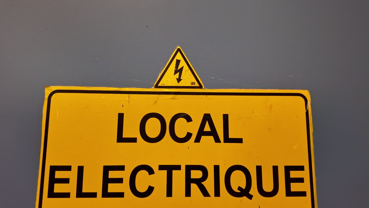 Close-up shot moving away from a yellow electrical hazard sign labeled "Local électrique" on a grey wall