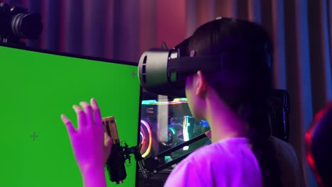 close up back view of asian girl streamer playing game, touching, and swiping experience virtual reality in futuristic goggles. green screen personal computer on desk illuminated by rgb led strip light