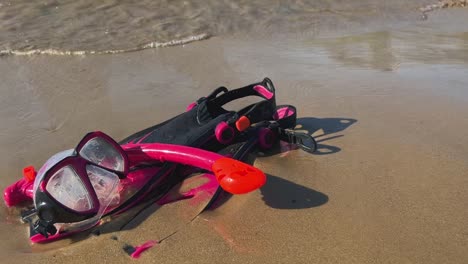 red and fuchsia fins mask and snorkel left on sandy beach with waves breaking on shore