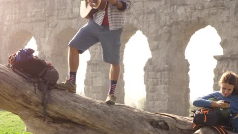 happy young couple backpackers tourists on a log trunk playing guitar singing reading book in front of ancient roman aqueduct ruins in romantic parco degli acquedotti park in rome at sunrise slow motion