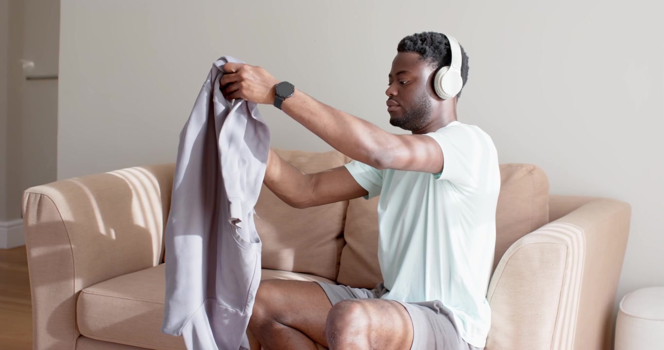 Erwachsener afrikanischer Mann trägt einen grauen Korb, betritt das Wohnzimmer, sitzt auf einem beigefarbenen Sofa und faltet Hemden.