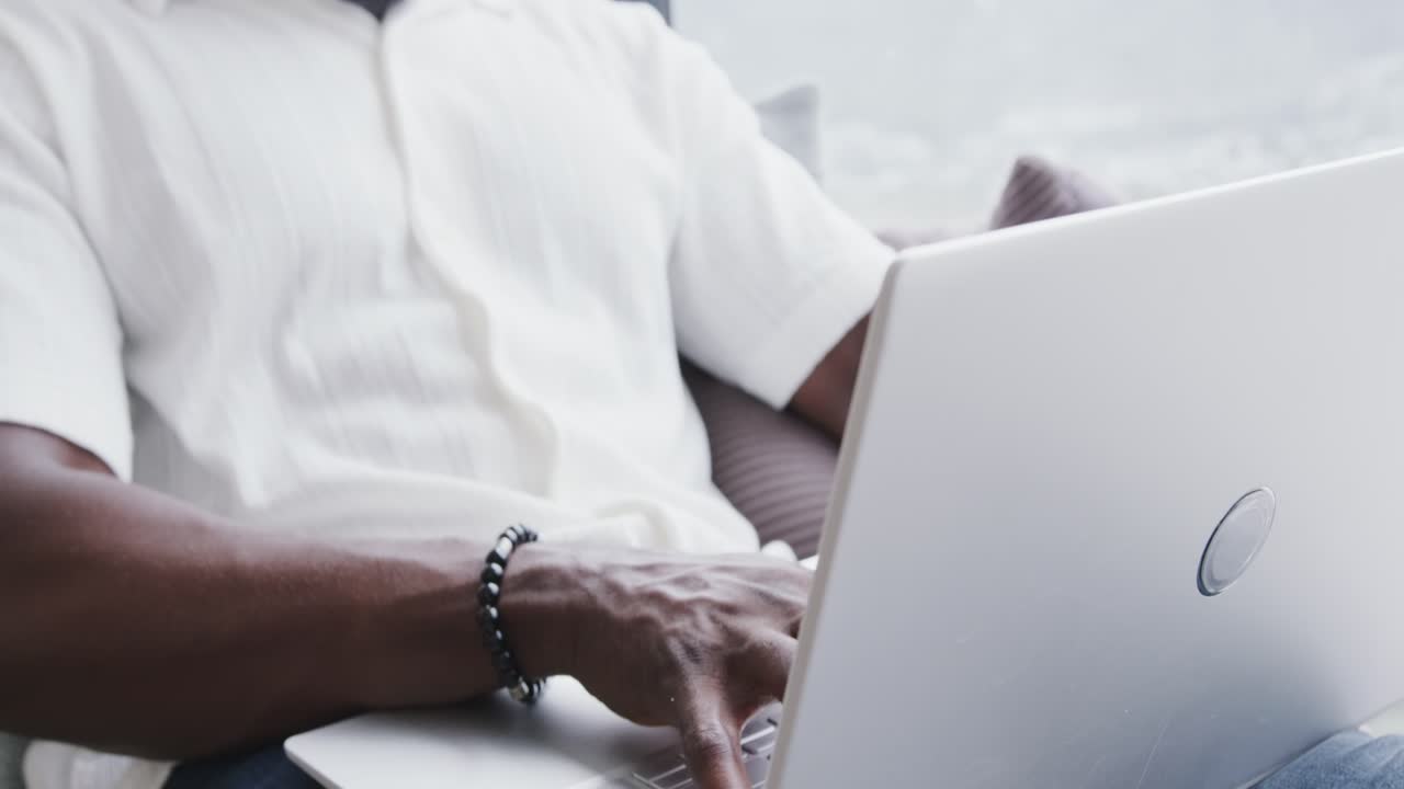 Uomo afroamericano che digita su un laptop bianco vicino a una grande finestra, indossando cuffie bianche e un braccialetto.