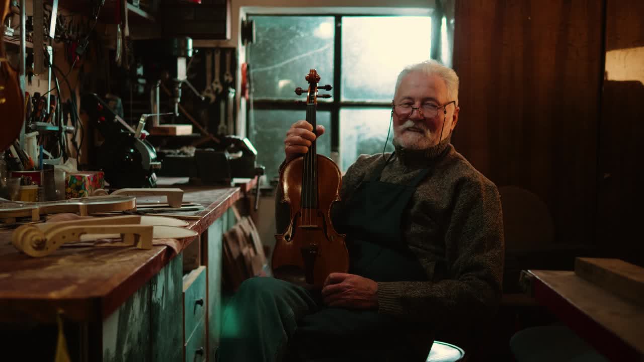 Senior professional violin master in his workshop smiles and looks at the camera