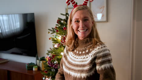 smiling woman wearing festive antlers decorating christmas tree at home