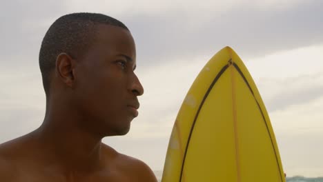 vista frontal de un surfista afroamericano de pie con una tabla de surf en la playa 4k