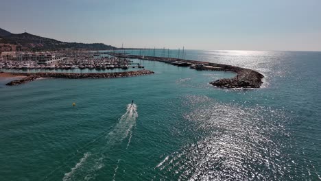 A-lone-skate-surfer-cruising-near-port-ginesta-in-barcelona,-spain,-glimmering-sea-under-the-sun,-aerial-view