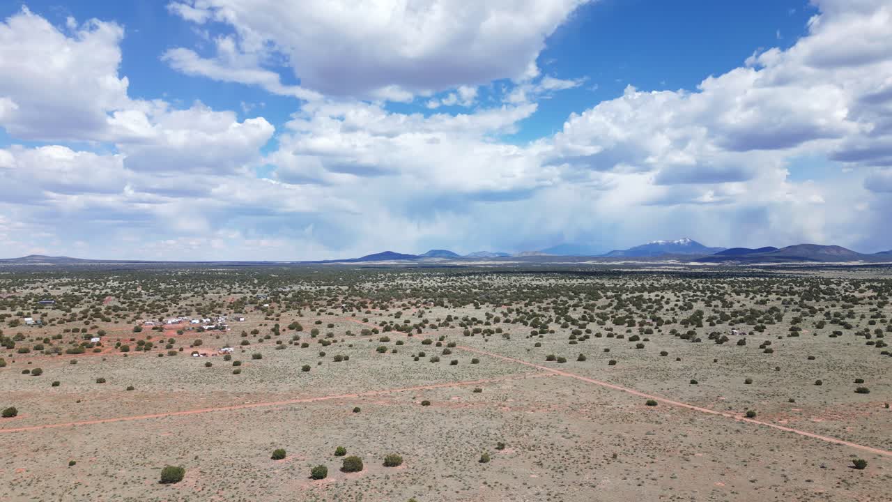 Drone rising over Williams, Arizona, south of the Grand Canyon. Clouds are dotting the sky over warm dirt on a bright sunny day. There are dirt roads, mountains and a few structures. Rain in the back