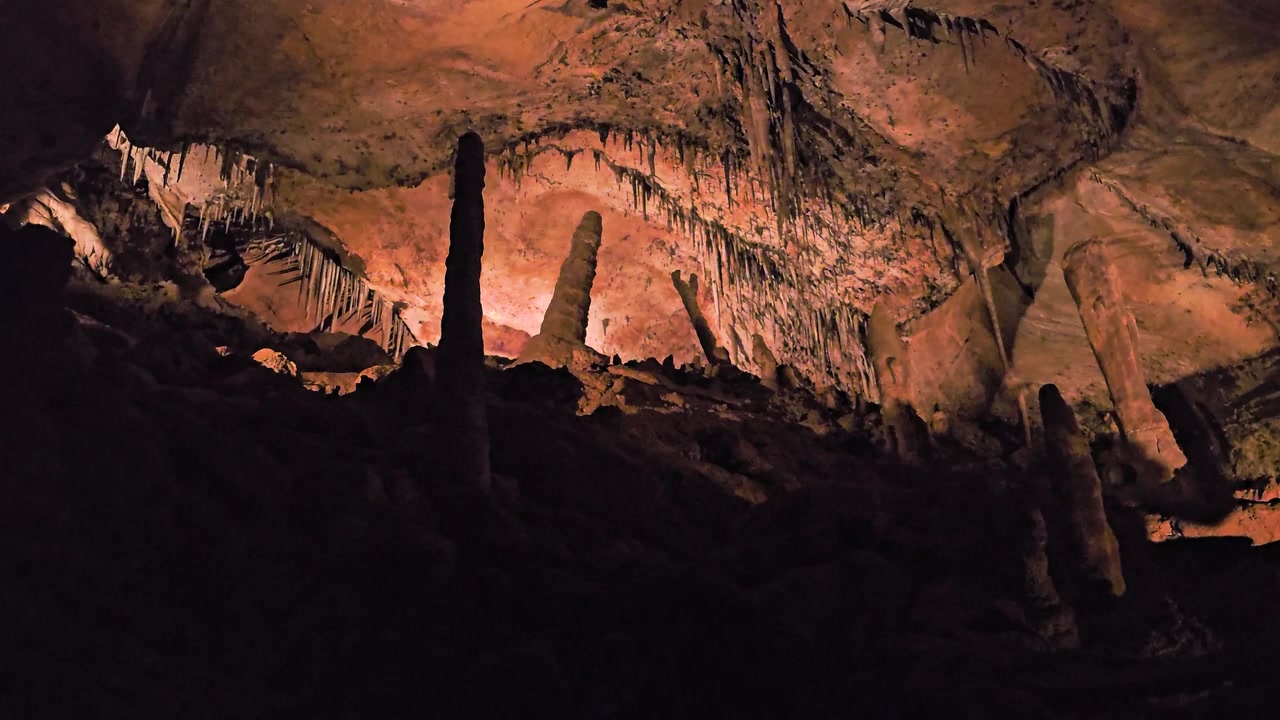 Dim limestone cave interior with stalactites, stalagmites, rocky floor, dramatic shadows, warm artificial lights, and textured mineral formations for underground nature footage.