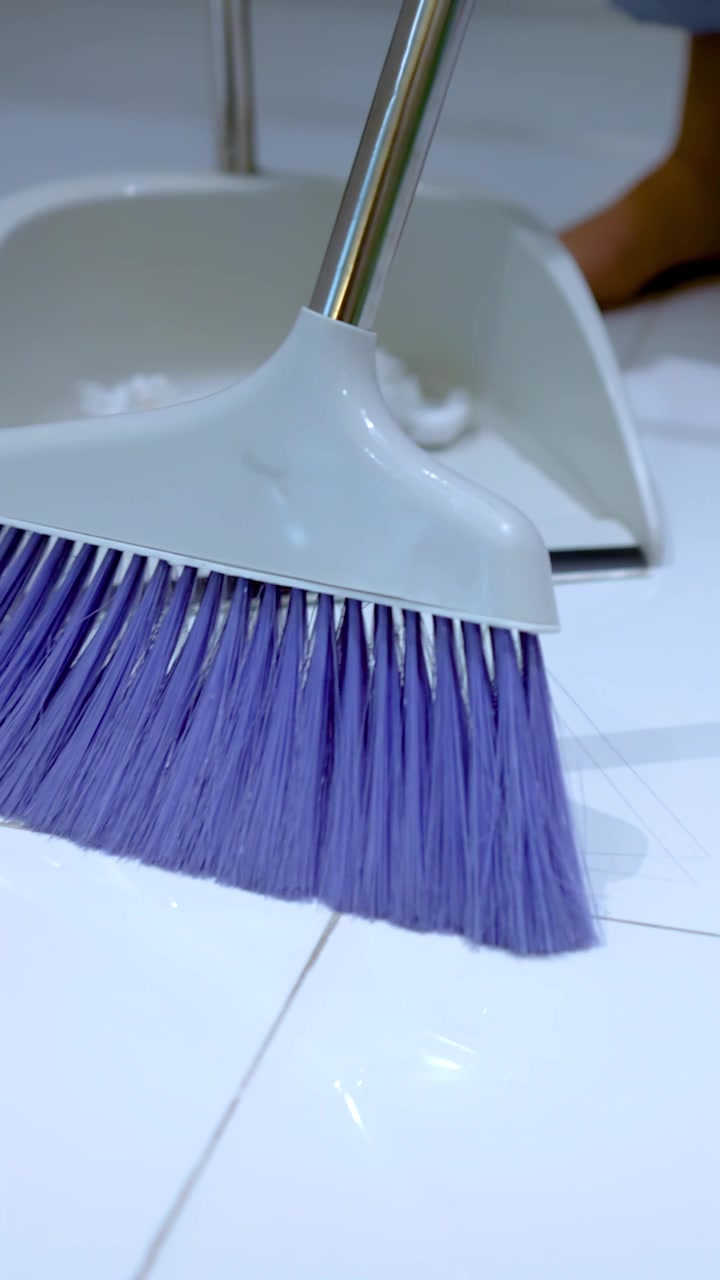 Close-up of bare feet and purple broom sweeping trash into a dustpan on tiles.