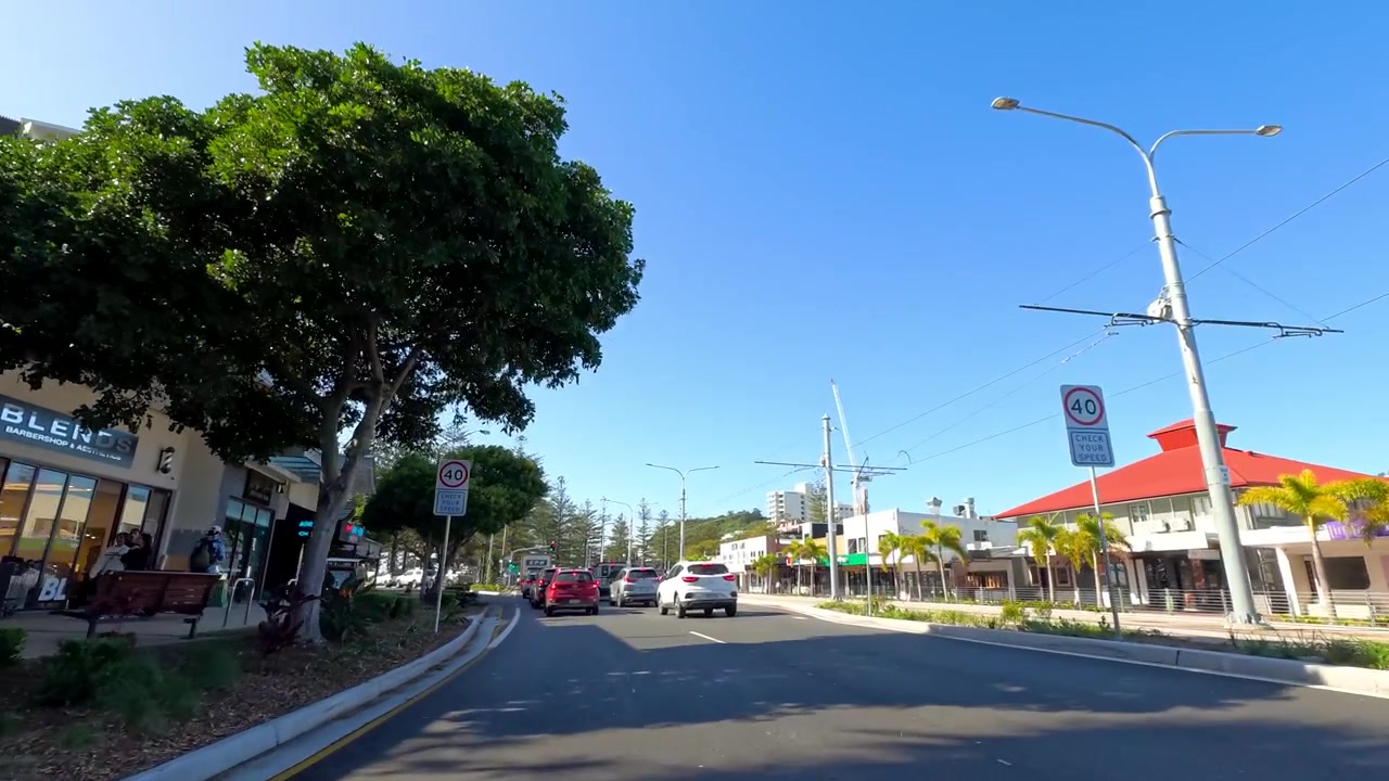 Une vue grand-angle depuis un véhicule parcourt une route côtière ensoleillée, passant devant des magasins et des appartements.