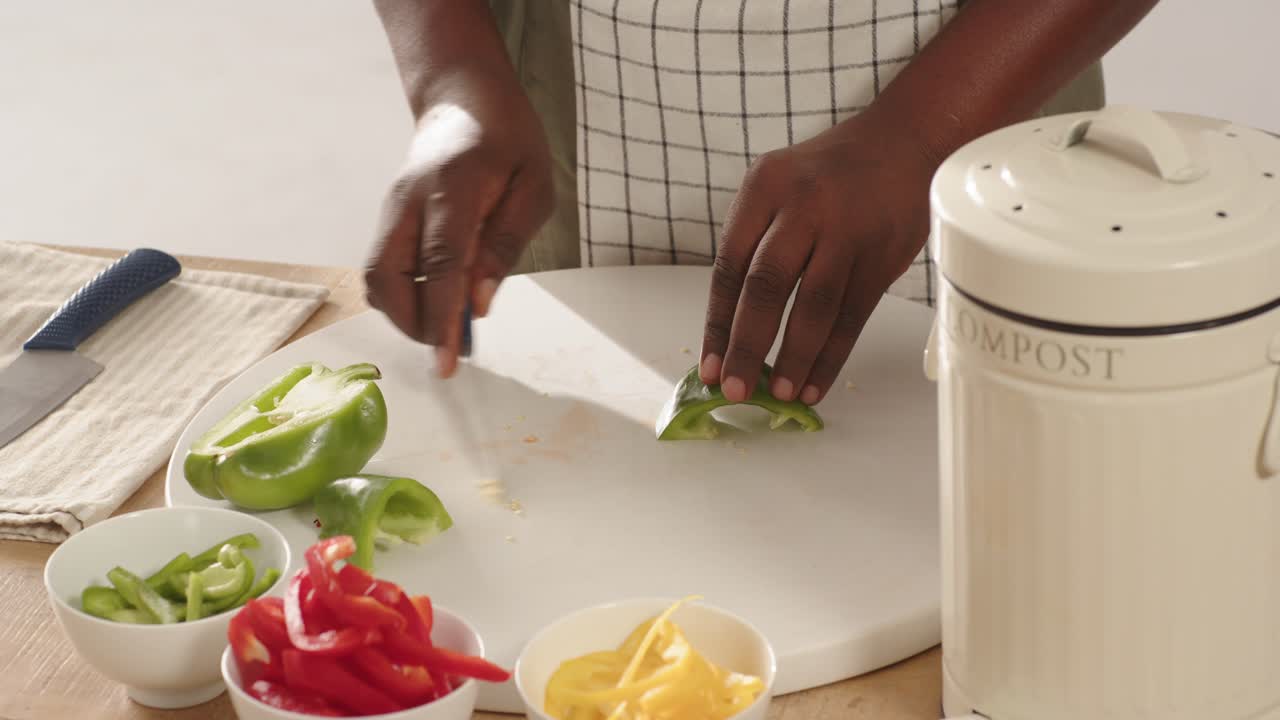 Homem afro-americano colocando metade de um pimentão verde no balcão, retirando o miolo para um balde de compostagem e fatiando para cozinhar.