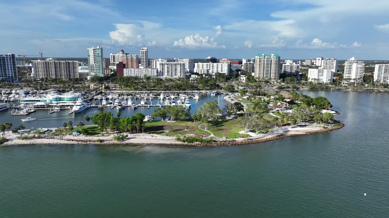 Harbor with yachts and sailing boats in Sarasota City, Florida. Aerial backwards wide shot. Sunny day with tropical Island and palm trees. High-rise Buildings and Apartment towers of Downtown.