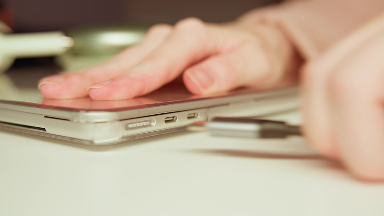 Close-up low-angle view shows slim laptop on white tabletop with black charging cable inserted at side port while hand presses nearby, creating visible flex, strain, and connector movement