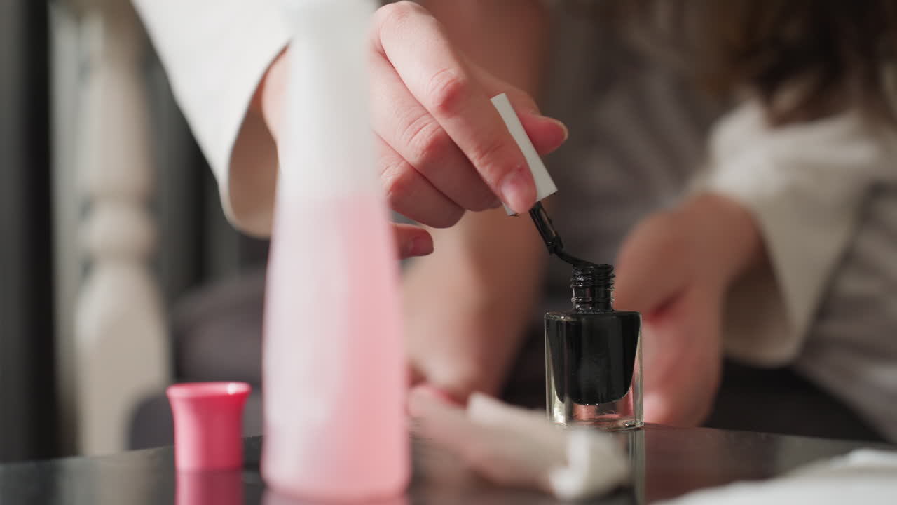 Black nail polish bottle on table beside pink remover and open cap, blurred female hands in background carefully brushing color onto toes, soft daylight, glossy surface reflection, quiet indoor mood
