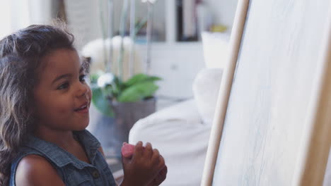 young hispanic girl drawing with pink chalk on a blackboard at home, close up, side view