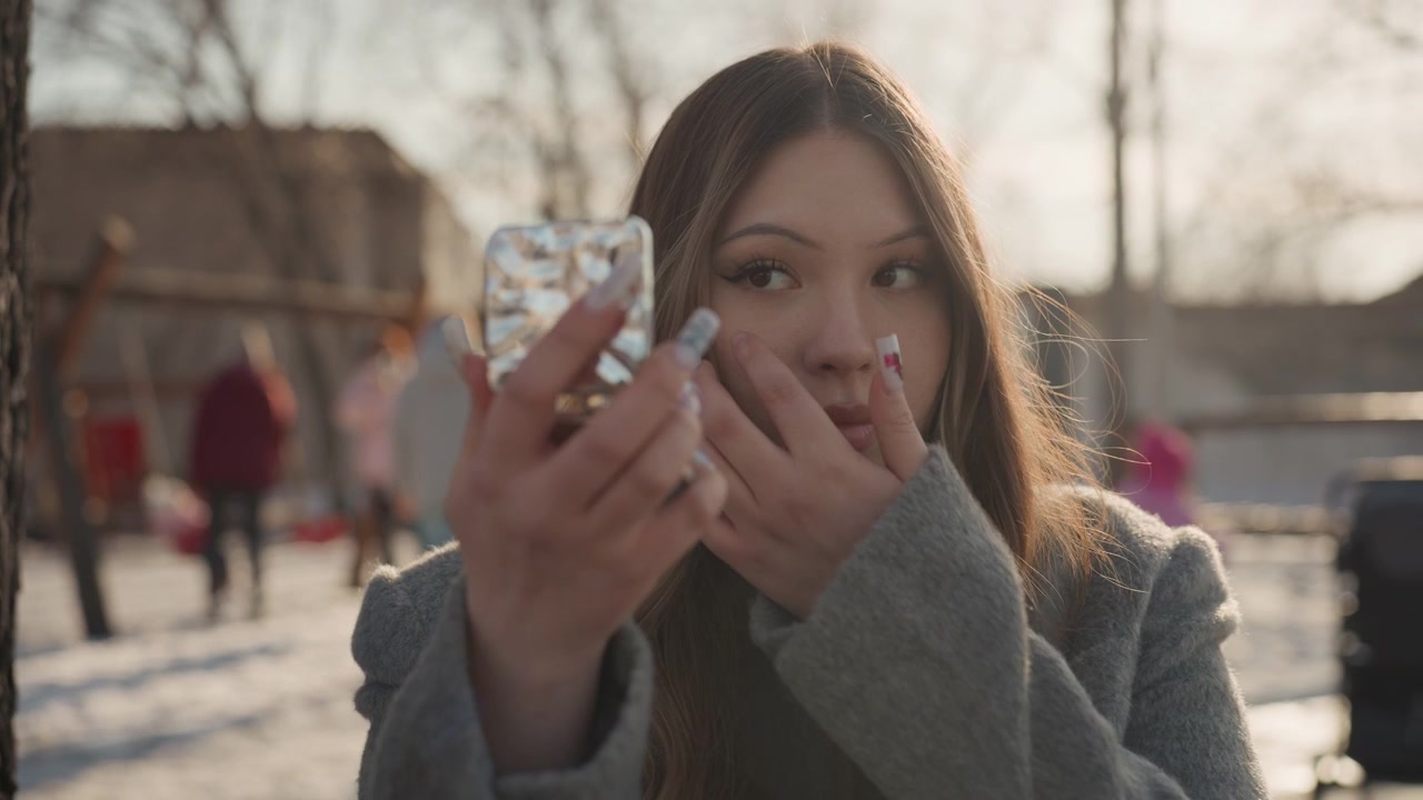 Bella donna asiatica in posa per la preparazione del trucco all'aperto in un parco invernale davanti a uno specchio, lunghi capelli retroilluminati, che applica il rossetto, energia da influencer/creatrice, passanti sullo sfondo, content creator che si prepara per una diretta streaming