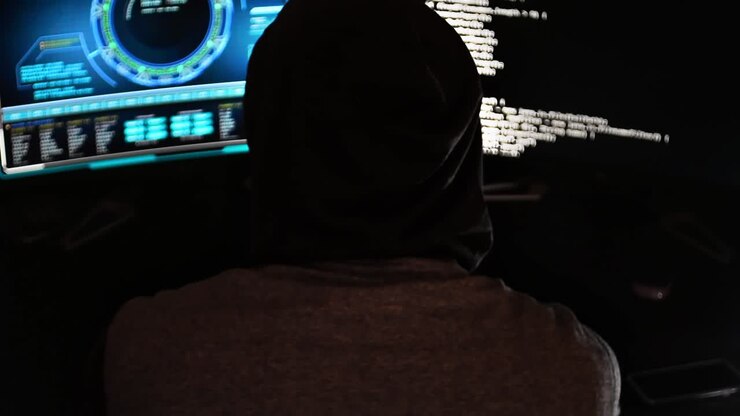 Inquadratura ampia di un hacker cybercriminale in un ufficio buio con schermi hi-tech