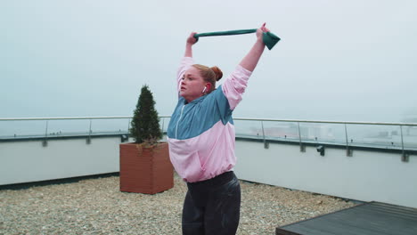 Sporty-girl-in-sportswear-does-yoga-stretching-exercising-using-rubber-band-on-roof-of-house-outdoor