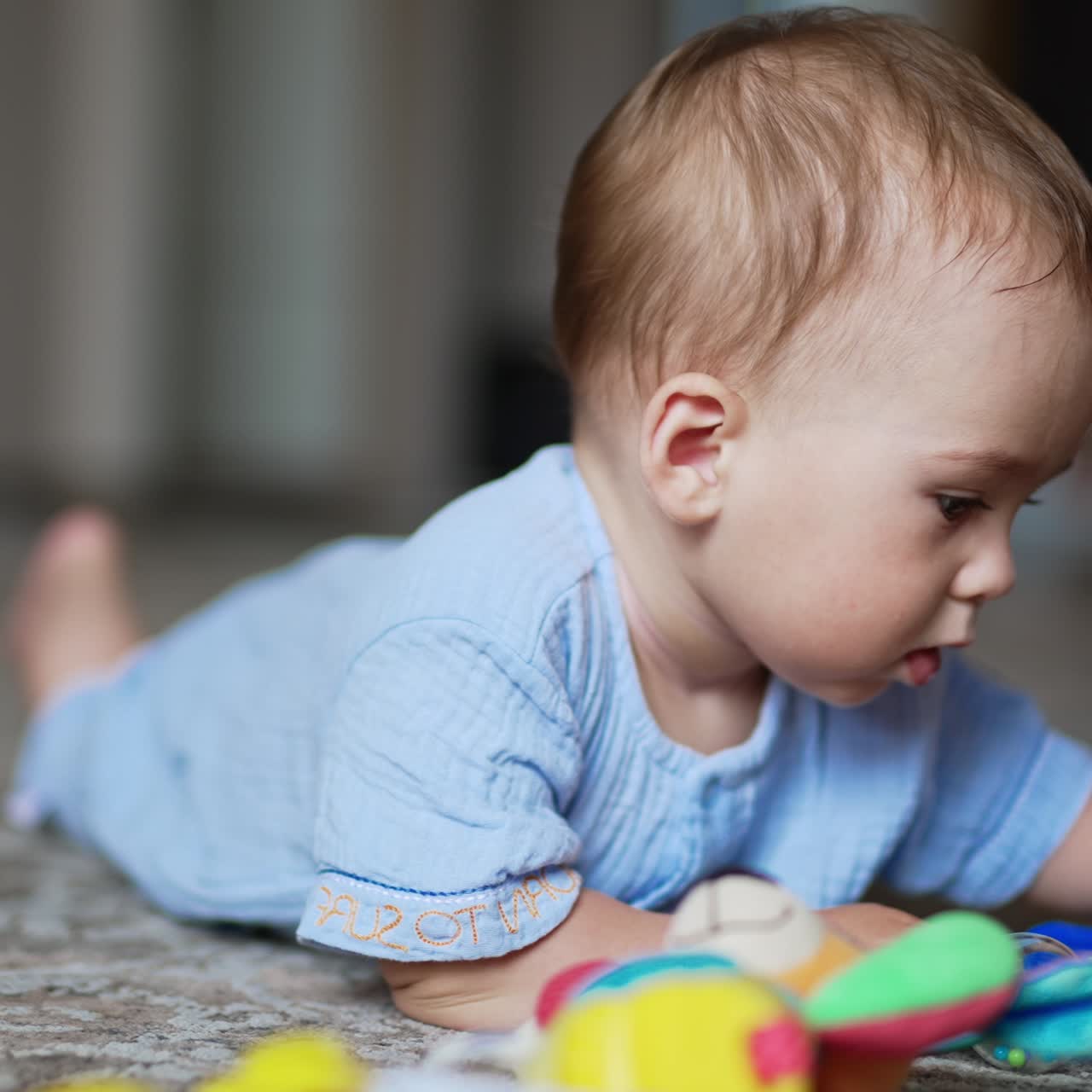Nice kid wearing blue shirt lies on the floor. Little boy touches toys lying beside him. Close up. Blurred backdrop