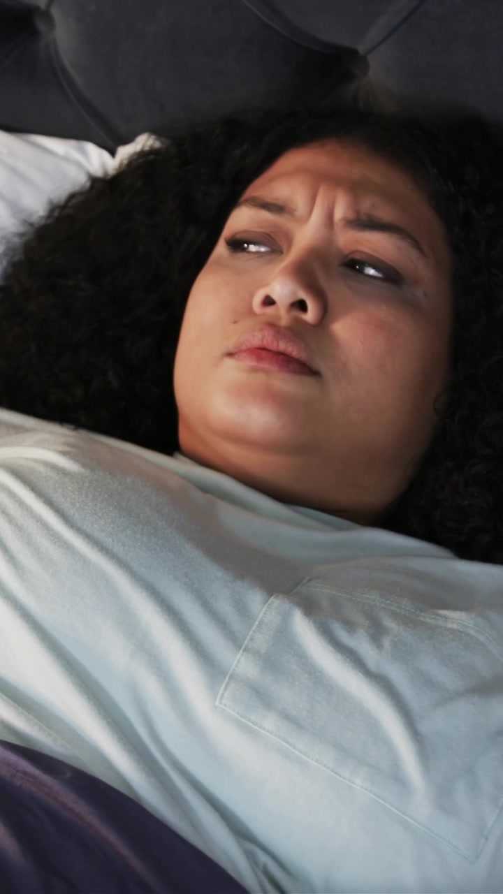 Vertical video: Waking woman in pale top turning, frowning in bed after sudden noise, purple cover