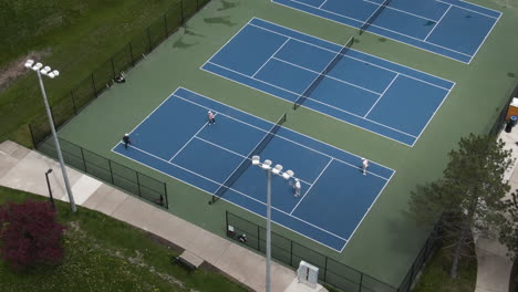 Un-Primer-Plano-Aéreo-De-Canchas-De-Tenis-Duras,-Donde-Los-Jugadores-Juegan-Dobles