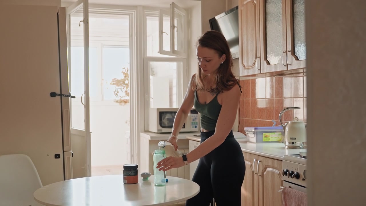 Donna riempie una bottiglia. Donna caucasica prepara l'idratazione a casa. Scena mattutina di donna che riempie una bottiglia in cucina. Donna in cucina riempie una bottiglia d'acqua per l'idratazione durante le ore mattutine.