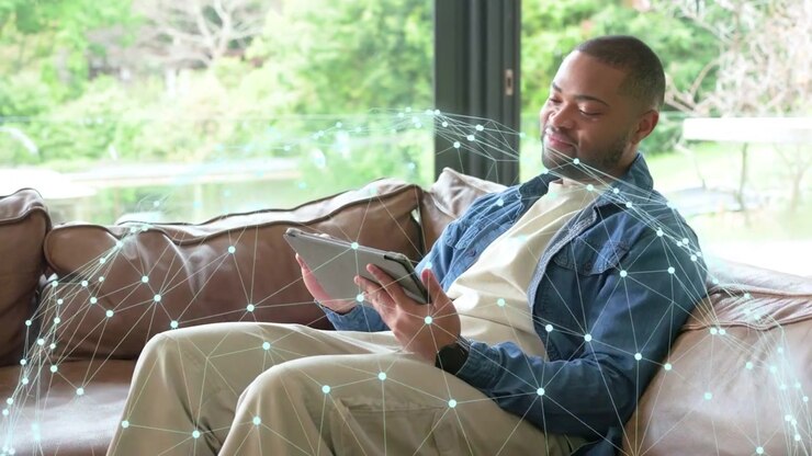 Man tapping tablet, scrolling and rotating while network overlay around sofa showing data for tech