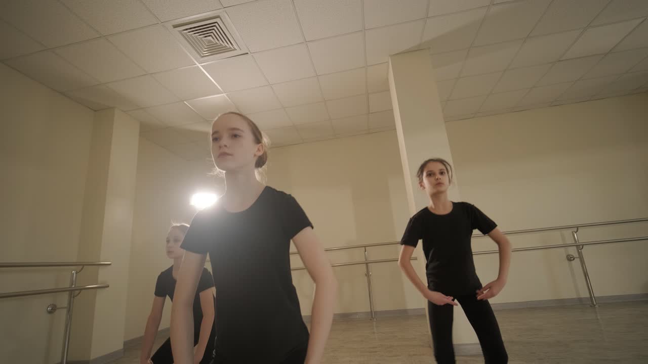A group of young ballet students in black dancewear practicing positions in a spacious ballet studio with wooden flooring and wall-mounted barres. Focused expressions and synchronized movements.