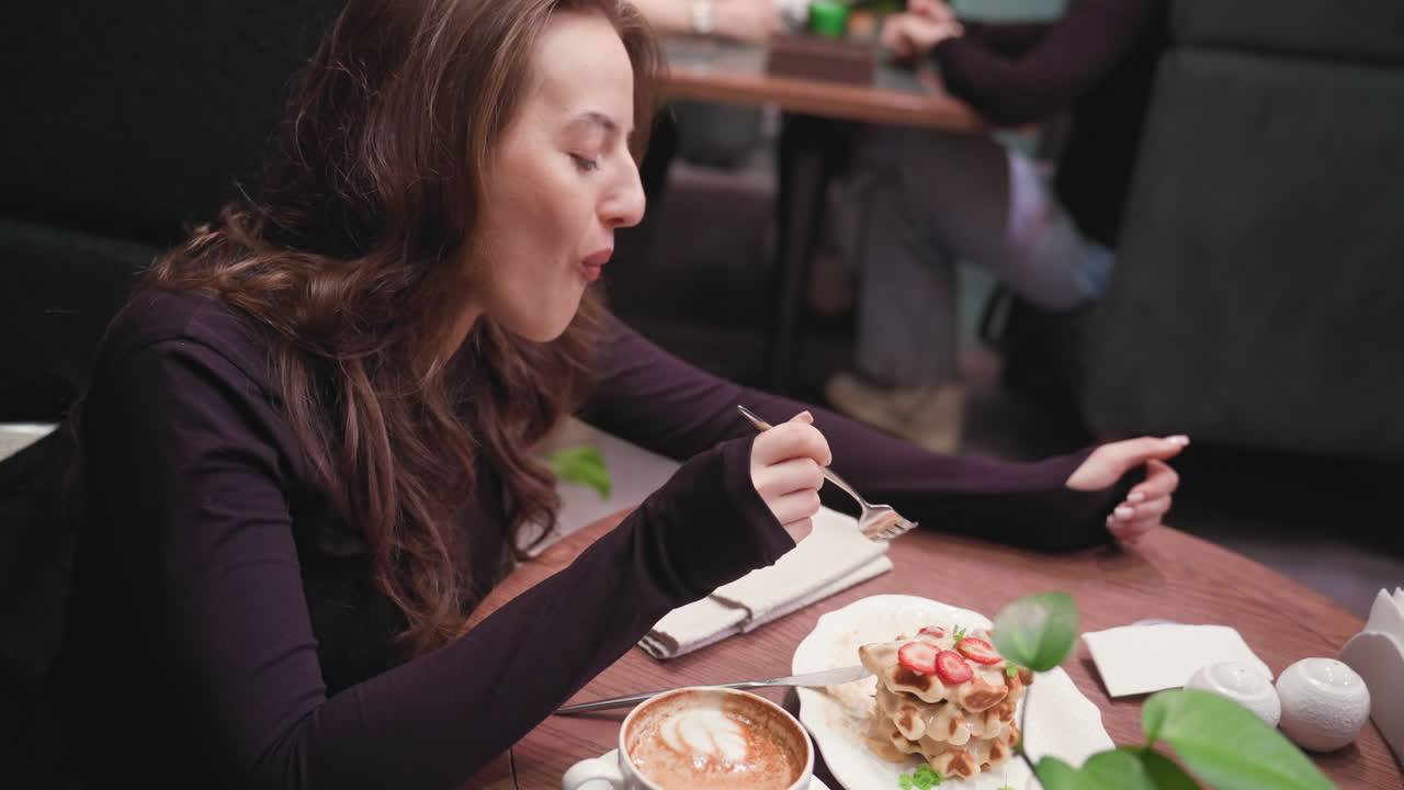 Lady enjoys delicious waffle stack topped with strawberries while seated at wooden table with cup of latte, as soft background blur shows other guests seated and engaged in casual conversation nearby