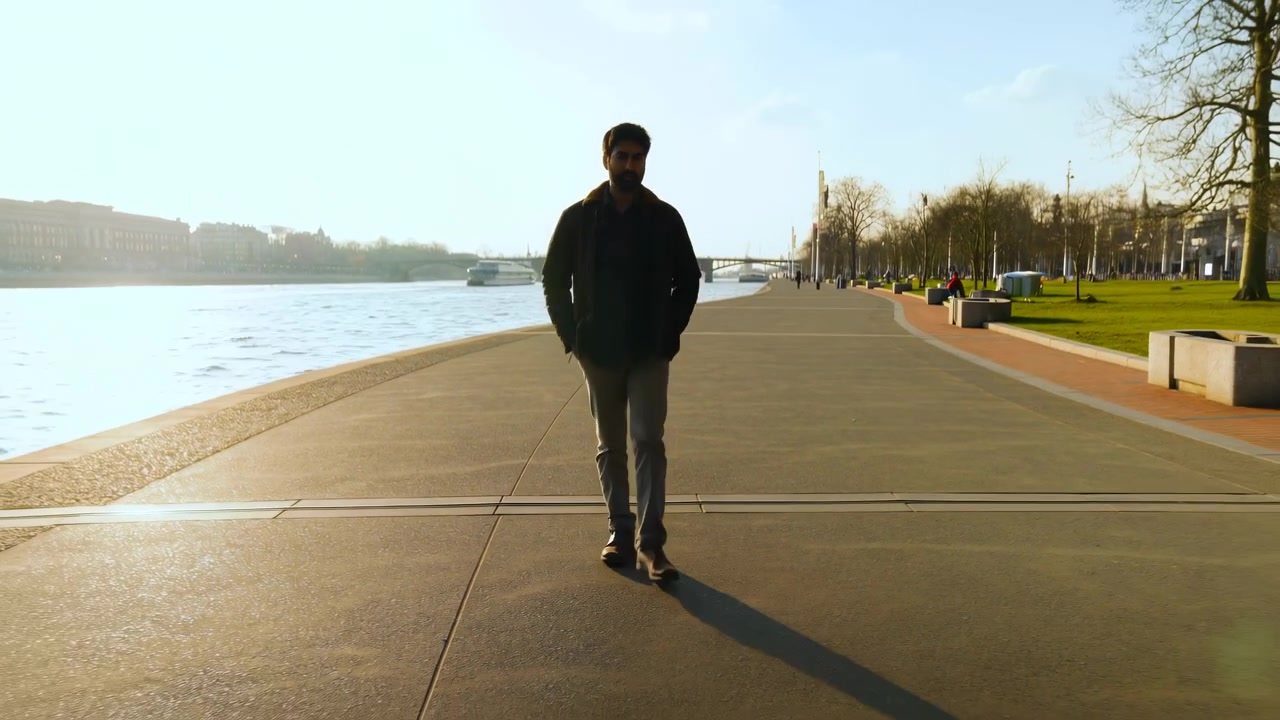 Un uomo adulto si avvicina costantemente su una passerella pavimentata sul lungomare sotto una calda luce solare bassa, incorniciato da acqua luminosa, barche e ponte in lontananza, alberi spogli, prato verde, sedute e un'ombra allungata in primo piano