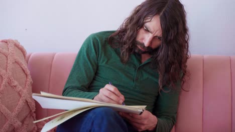 plano medio del artista barbudo hispano con el pelo castaño largo en camisa verde y jeans azules sentado en un sofá rosa dibujando diligentemente en el cuaderno de dibujo