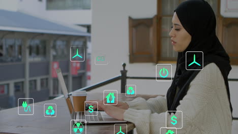 Animation-of-multiple-icons-over-biracial-woman-wearing-hijab-working-on-laptop-with-coffee-cup