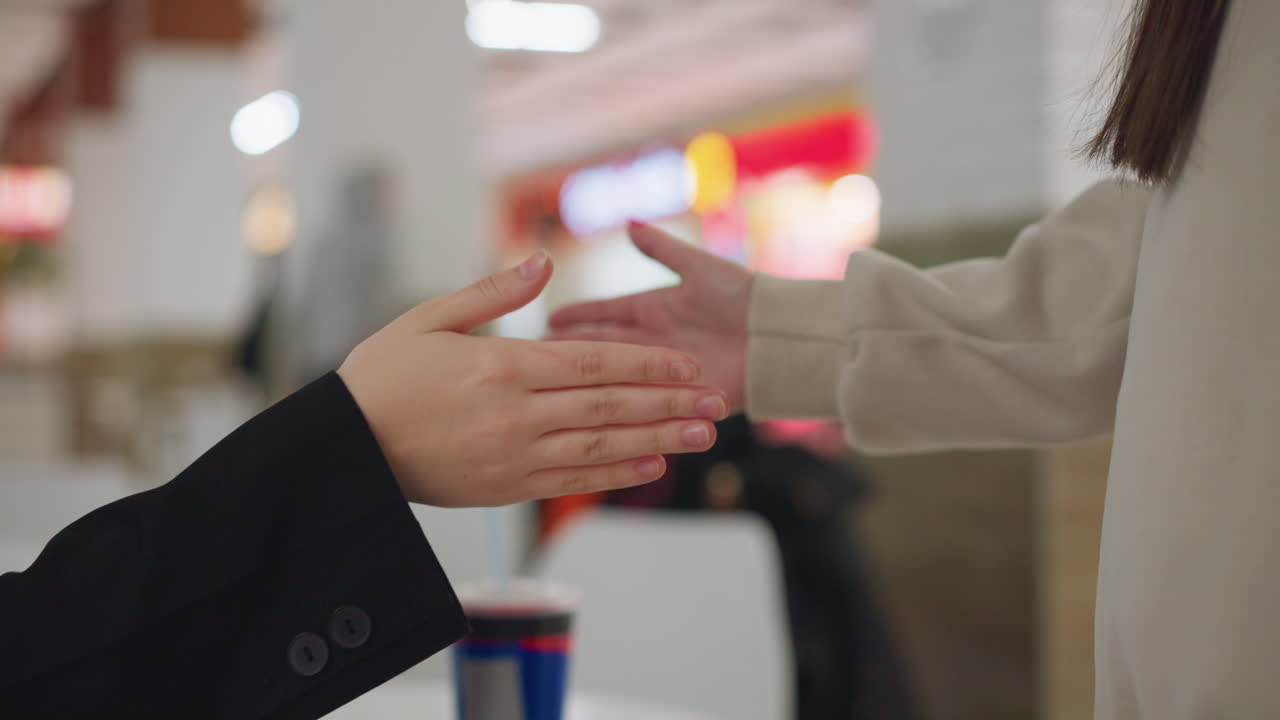 Close up of two ladies greeting in funny way with extended hands, capturing playful moment of friendship and connection in casual indoor setting with blurred people and lights in background