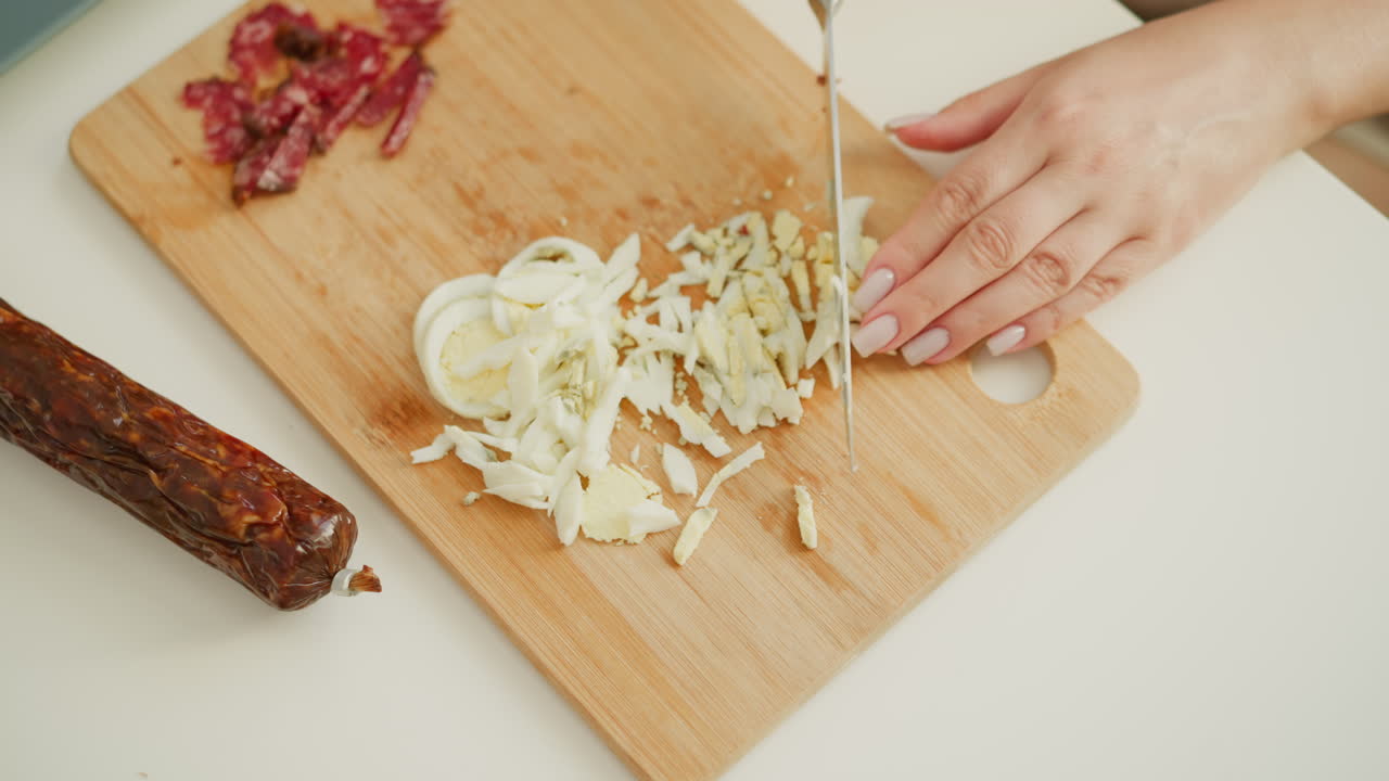 Hand of female baker carefully cutting boiled egg into tiny pieces beside sliced sausage on wooden board, showcasing precise kitchen skills and neat food preparation in bright casual kitchen setting