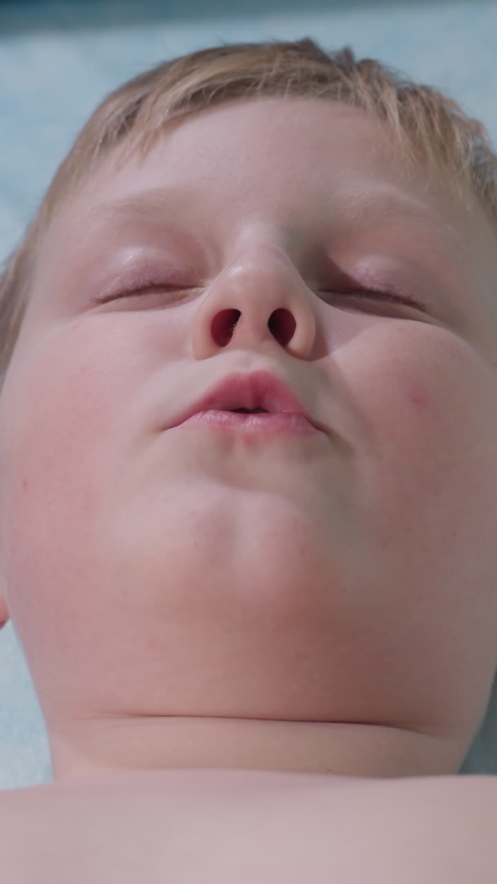 Close de um menino branco deitado de costas, respirando lentamente durante um exercício de relaxamento, iluminação suave e fria na pele, sardas sutis na bochecha, olhos fitando para cima e depois se fechando, lábios se apertando na expiração, terapia pediátrica