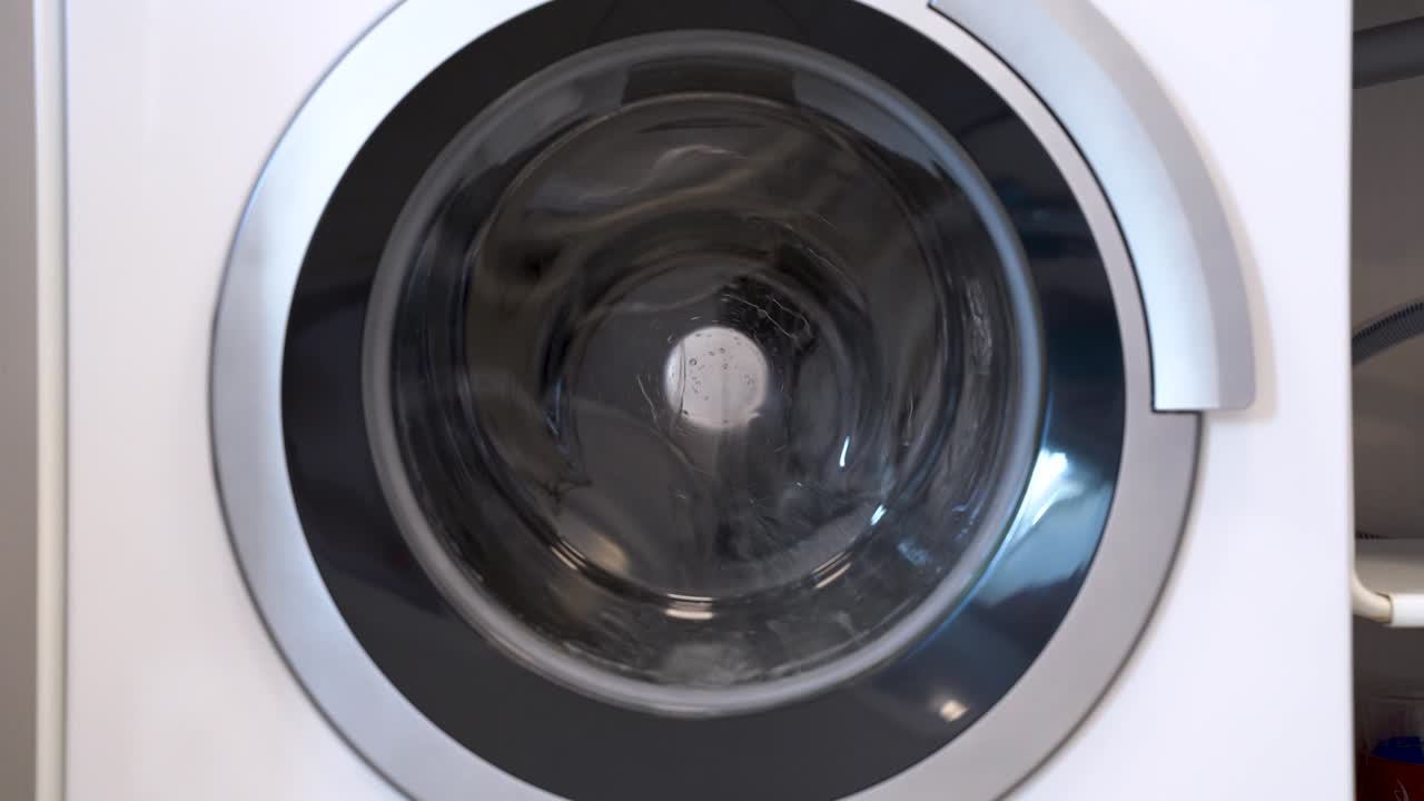 Close up shooting of washing machine drum, beginning to rotate, foam from detergent flows down the front glass. Indoors, white background.