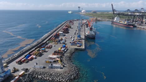 Aerial-flight-over-jetty-at-port-of-Caucedo-with-crane-and-goods-during-sunny-day---Oil-polluting-Caribbean-Sea---Environmental-pollution-at-industrial-harbor