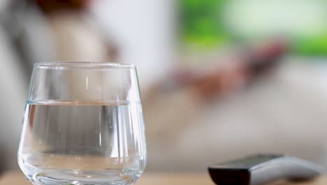 a clear glass of water on a table vibrates gently, with a blurred figure holding a remote in a bright, modern living room