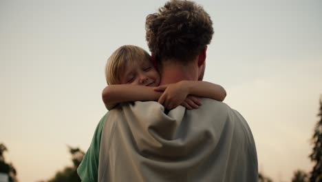 A-little-blond-boy-lovingly-hugs-the-neck-of-his-dad-with-curly-hair-in-a-green-T-shirt.-Little-boy-loves-his-dad-and-hugs-him