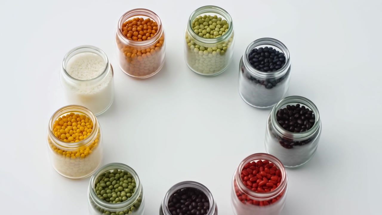 Vista dall'alto ad angolo elevato di barattoli di vetro trasparenti aperti disposti in un anello circolare uniforme su un tavolo bianco pulito, ogni barattolo riempito con piccoli pellet uniformi in colori saturi, creando una geometria ordinata.