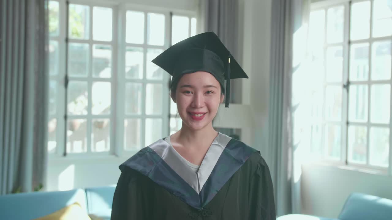 Asian Woman Wearing A Graduation Gown And Cap Smiling To Camera In Living Room