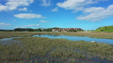 un dron de bajo vuelo disparó a través de los pantanos, revelando un reflejo en el agua del cielo azul y las nubes esponjosas, luego continúa hacia un velero y el pueblo de alnmouth, northumbria, inglaterra.