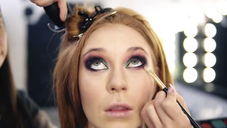 front footage of a blonde beautiful model sitting and looking up while make up artist finishing her smoky eyes . hairdresser behind