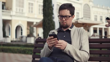 serious man with glasses prints sms on a smartphone in the park on a bench