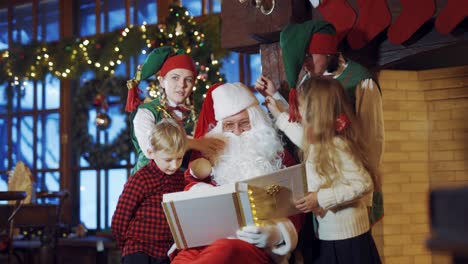 children and elves touch santa's white beard and red cap on christmas background. santa claus is sitting with album and having fun with two little kids and fairy elves indoors.