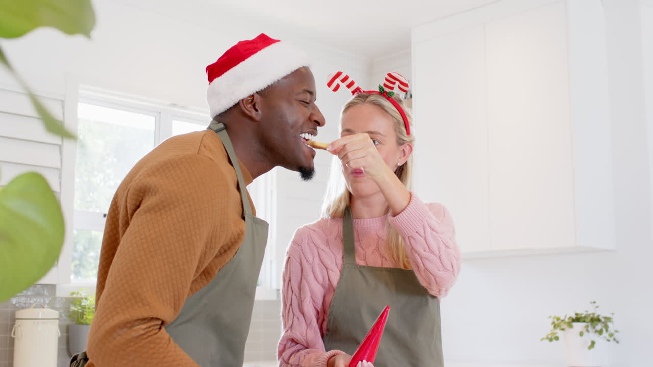 Couple decorating Christmas cookies together, wearing festive hats in kitchen
